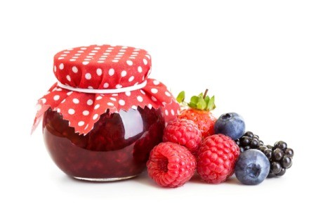 A jelly jar full of red jam with a red and white cloth on top. and fresh raspberries, blueberries, and blackberries beside it.