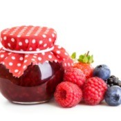 A jelly jar full of red jam with a red and white cloth on top. and fresh raspberries, blueberries, and blackberries beside it.