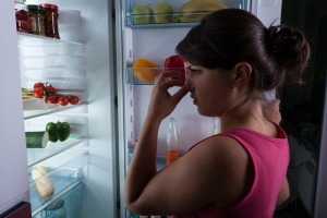 Woman holding nose while standing in front of open refrigerator door