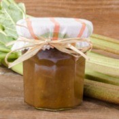 Jar of Rhubarb Marmalade with cloth covered lid in front of several stalks of rhubarb