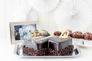 Double heart shaped chocolate cake that says "Wedding Anniversary" with a 25 decoration.  Black and white wedding picture in the background