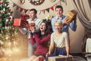 Two men and two women laughing and shaking presents by a Christmas tree.