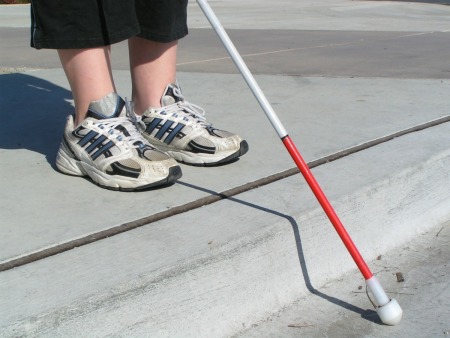 Woman's feet in sneakers near a curb holding a while cane with red marking.