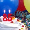Cake with candles that say 85 surrounded by balloons, ribbons, and confetti