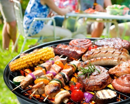 Barbecue grill with a variety of meats, shish kabobs, corn and veggies with a family in the background