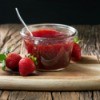 Container of strawberry jam with spoon and loose strawberries in front