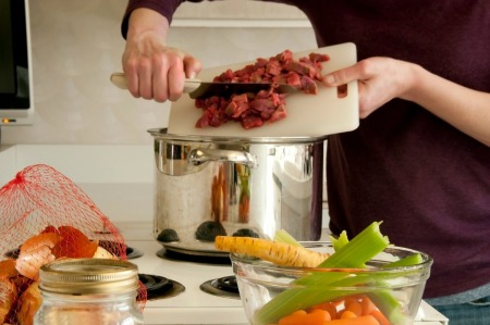 Woman pushing cubes of beef from cutting board into a soup pot with vegetables in the foreground.