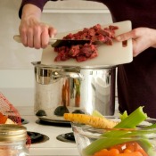 Woman pushing cubes of beef from cutting board into a soup pot with vegetables in the foreground.