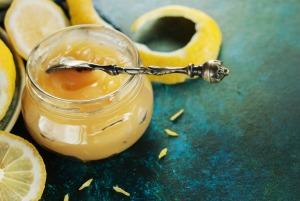 Open jar of lemon curd with spoon surrounded by lemons and lemon peel
