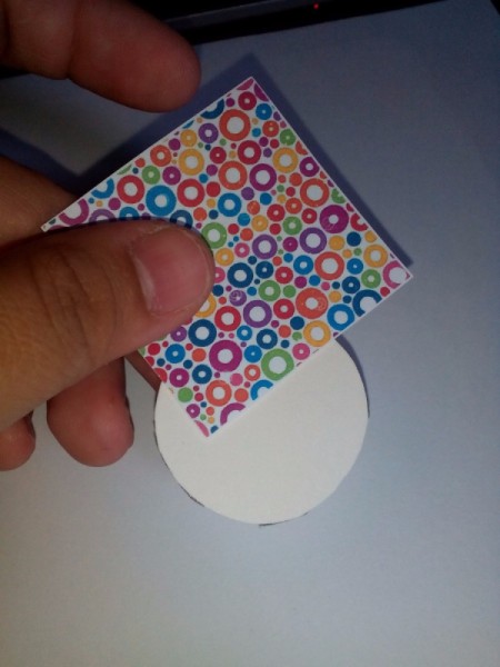 Hand holding a small square of the colored paper over the full circle frame backing ready to glue together.