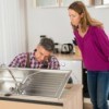 Woman standing over kitchen sink as man is reaching under to fix it.