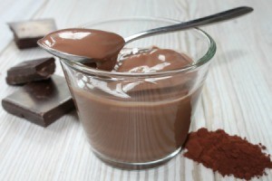 Glass dish of chocolate pudding with spoon surrounded by cocoa powder and pieces of chocolate bar