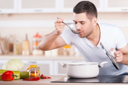 Man tasting from a pot of soup.  There are a variety of vegetables and peppers on the counter