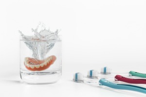 Dentures dropping into water glass with a splash, surrounded by three toothbrushes