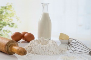Flour, milk, eggs, butter, rolling pin and whisk on a counter top.