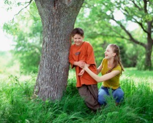 Boy and girl laughing and playing next to a tree trunk surrounded by tall weed-type grass