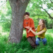 Boy and girl laughing and playing next to a tree trunk surrounded by tall weed-type grass