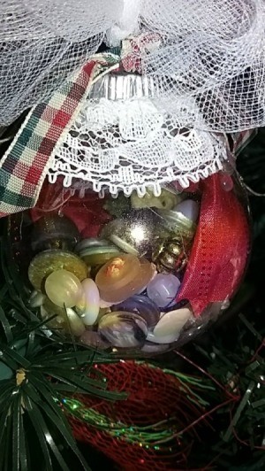 Clear plastic ornament filled with buttons and decorated with lace, netting, and ribbon hanging on the tree.