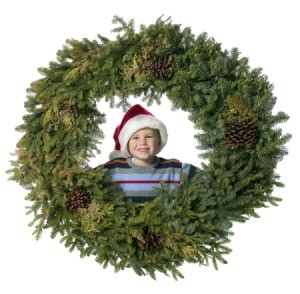 A boy smiling looking through the opening of a giant Christmas wreath.