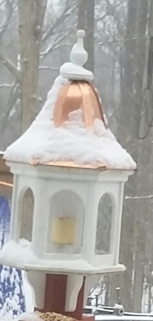 A white birdfeeder with a copper roof and a pillar candle inside where the birdfood would normally go.