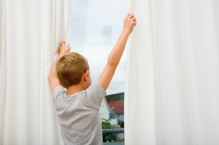 A boy holding onto white curtains as he looks out the window.