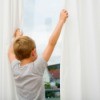 A boy holding onto white curtains as he looks out the window.