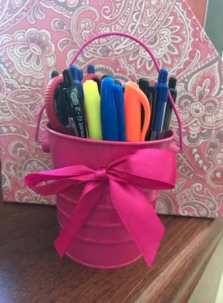 A pink tin pail filled with pens and markers on a desk.