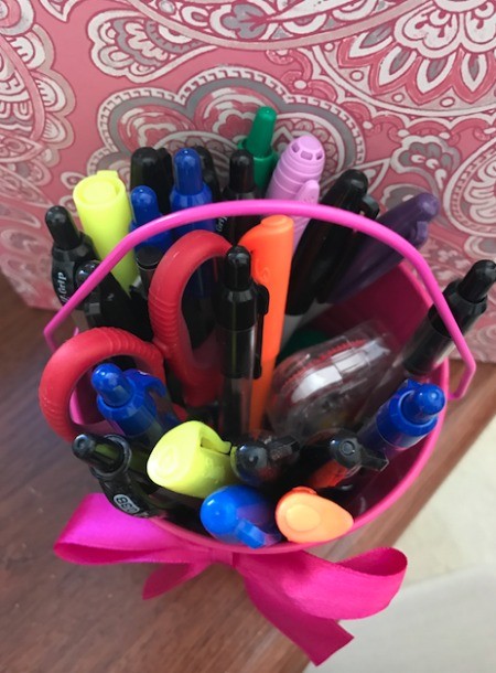 A pink tin pail filled with pens and markers on a desk.