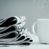 A stack of newspaper next to a cup of coffee.