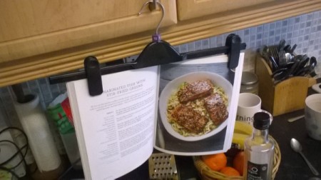 A pants hanger being used to display a cookbook in the kitchen.