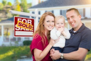 A happy couple with a baby outside a home with a for sale sign, marked sold.