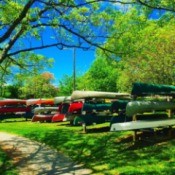 A collection of kayaks, canoes and other boats stacked on a green lawn.