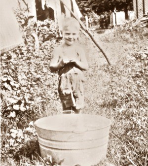 A sepia photograph of a young boy with a metal tub outside.