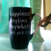 A mug that says "Happiness anytime anywhere, just pick it up" next to a window and a green vase.