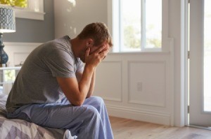 A depressed man with his face in his hand sitting on the edge of his bed.