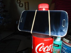 A soda bottle and rubber band being used as a phone stand.