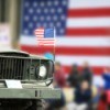 Close up of a Jeep with American flag flying on the hood and an American flag in the background.