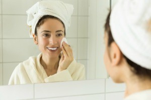 Woman looking in the mirror with a towel on her head using a cotton pad on her face.