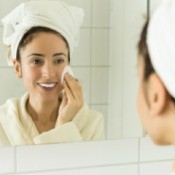 Woman looking in the mirror with a towel on her head using a cotton pad on her face.