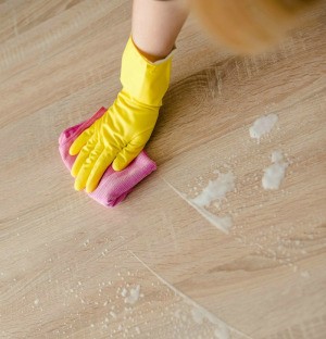 A gloved hand holding a sponge and soapy water on a wood surface.