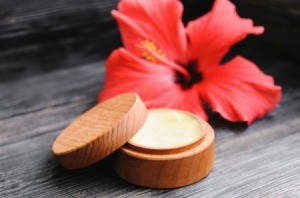 A wooden jar filled with lip balm and a pink flower on a wooden background.