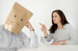 Abusive wife pointing at a man with a paper bag on his head with a sad face.