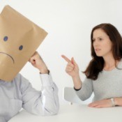 Abusive wife pointing at a man with a paper bag on his head with a sad face.