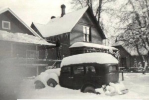 A black and white photo of a house with snow on the roof and on the old fashioned car in front.