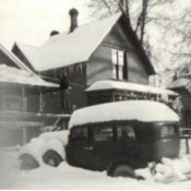A black and white photo of a house with snow on the roof and on the old fashioned car in front.