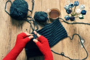 Person wearing red fingerless gloves knitting something grey.