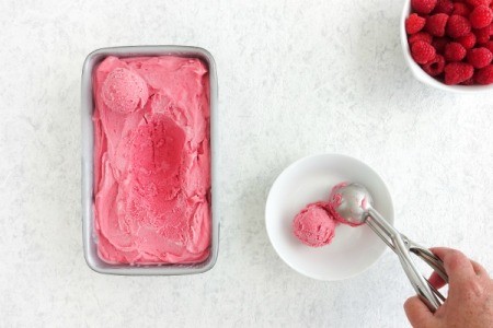 Person scooping from a tub of ice cream next to a bowl of fresh raspberries.