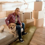 Man sitting on a plastic covered sofa with his dog, surrounded by boxes.