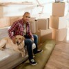 Man sitting on a plastic covered sofa with his dog, surrounded by boxes.