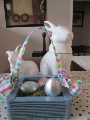 Craft Stick Basket  - blue basket with spring colored checkered ribbon handle and pearlized eggs inside, sitting in front of two white ceramic rabbits